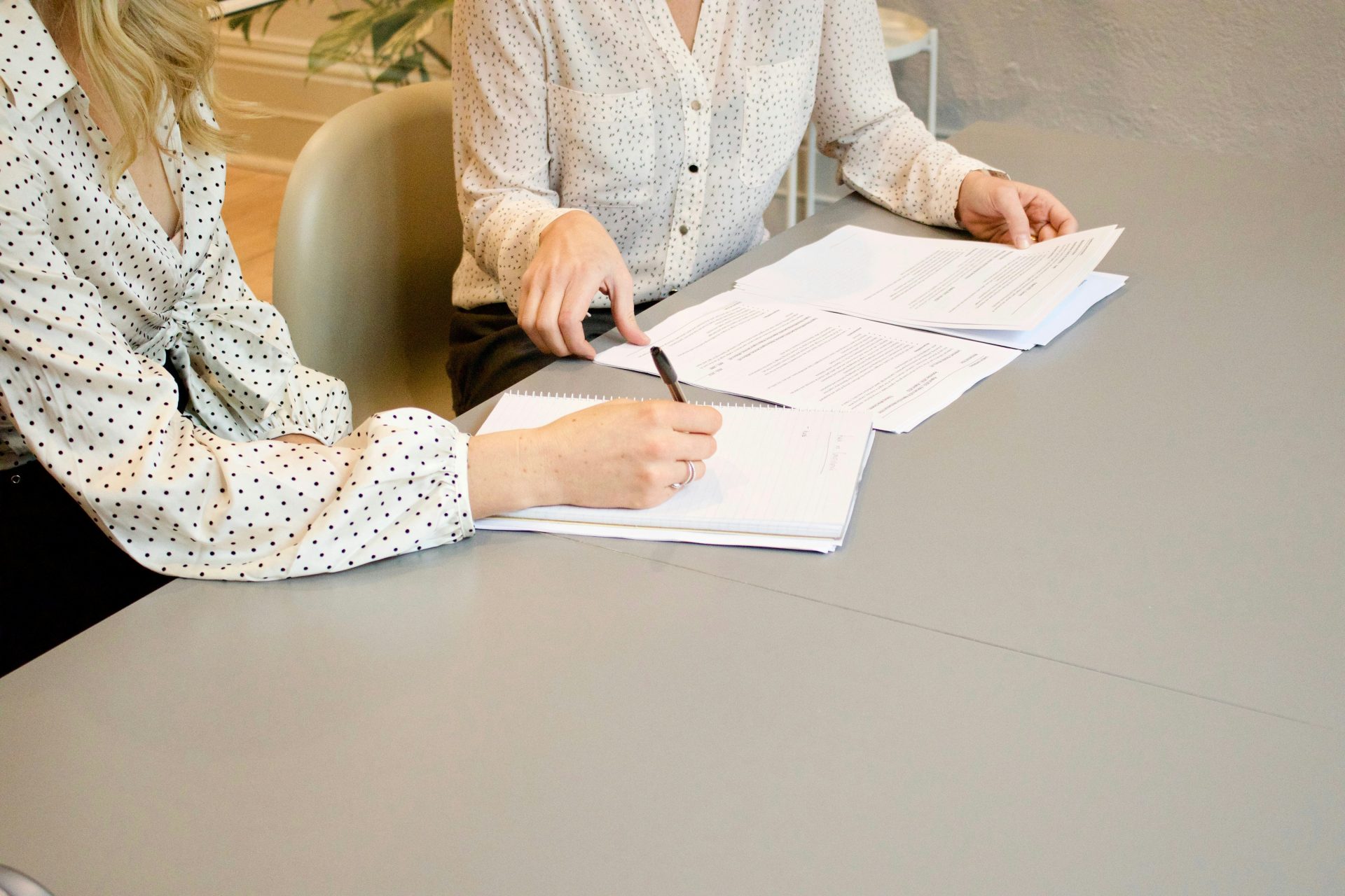 Une femme signe sur une feuille de papier blanc d'imprimante à côté d'une autre femme qui s'apprête à toucher les documents.